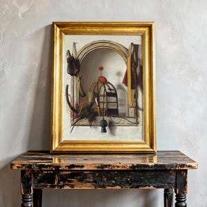A framed painting with a gold border depicts a whimsical still life with a cage, various tools, and objects against an arched backdrop, resting on a rustic wooden table. The scene feels nostalgic and surreal.