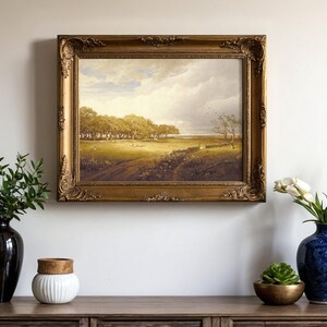 A framed landscape painting is centered on a wall, depicting a serene countryside scene. Below, a wooden console holds vases with flowers and greenery.