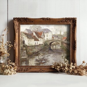 A rustic framed painting of a serene village scene with a bridge over a river, flanked by houses. Dried flowers in jars enhance the cozy atmosphere.