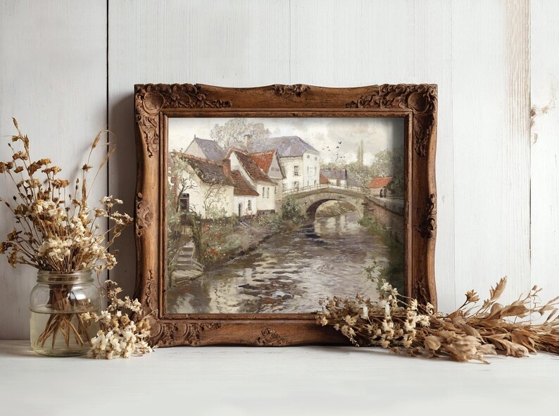 A rustic framed painting of a serene village scene with a bridge over a river, flanked by houses. Dried flowers in jars enhance the cozy atmosphere.