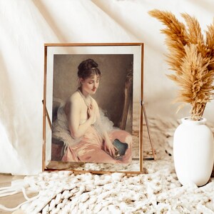 Framed portrait of a woman in a pink dress sits on a fluffy white rug. Beside it, a white vase holds dried pampas grass, creating a serene, elegant scene.
