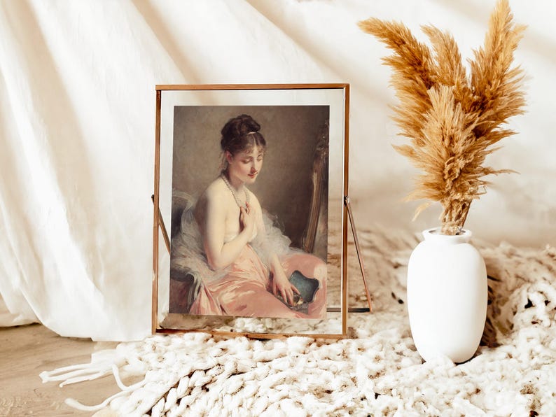 Framed portrait of a woman in a pink dress sits on a fluffy white rug. Beside it, a white vase holds dried pampas grass, creating a serene, elegant scene.