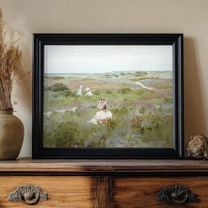A framed painting on a wooden sideboard depicts women in a serene meadow. Nearby, a vase with pampas grass adds a rustic touch. Calm, pastoral scene.