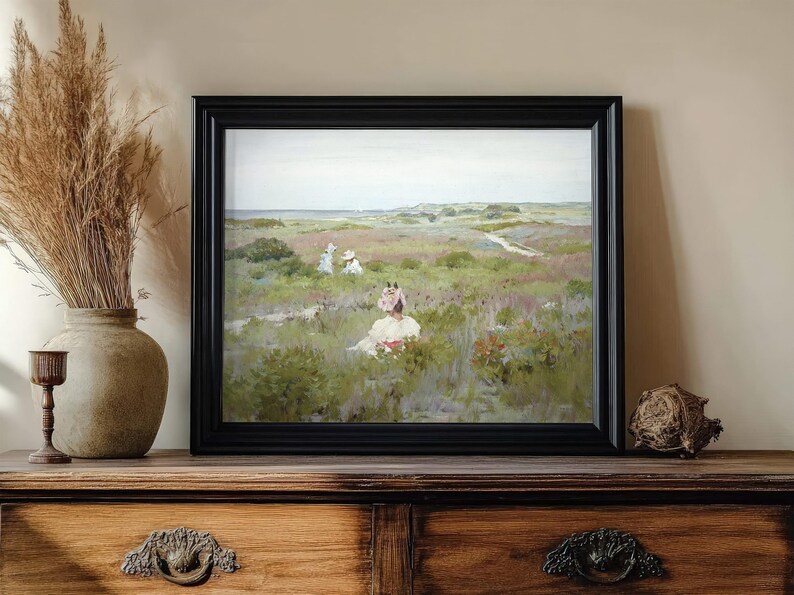 A framed painting on a wooden sideboard depicts women in a serene meadow. Nearby, a vase with pampas grass adds a rustic touch. Calm, pastoral scene.