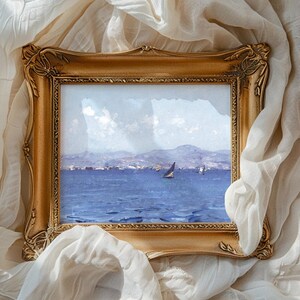 An ornate golden frame encases a seascape painting with a sailboat on blue waters, set against distant mountains, resting on white fabric.