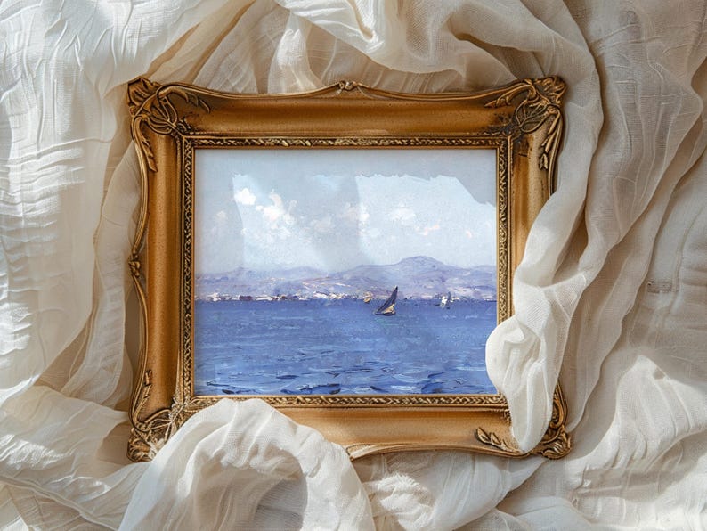 An ornate golden frame encases a seascape painting with a sailboat on blue waters, set against distant mountains, resting on white fabric.