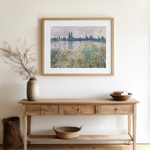 A serene living space with a wooden console table, a framed impressionist landscape painting above it, a vase with dried branches, and a bowl on the table.
