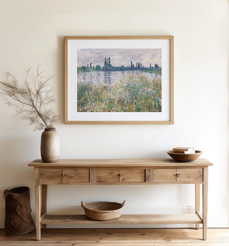 A serene living space with a wooden console table, a framed impressionist landscape painting above it, a vase with dried branches, and a bowl on the table.