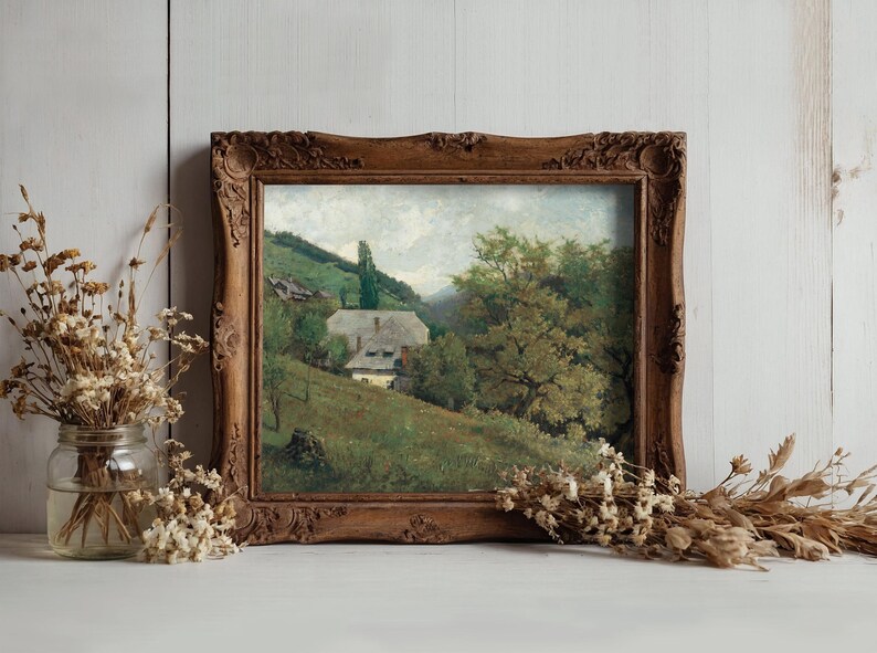 A vintage painting in an ornate wooden frame depicts a serene countryside with a house, trees, and hills. Nearby, dried flowers in a glass jar enhance the rustic ambiance.