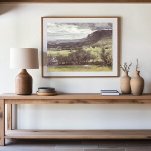 A serene living room setting with a wooden table holding a lamp, ceramic vases, and books, beneath a framed landscape painting of mountains and fields.