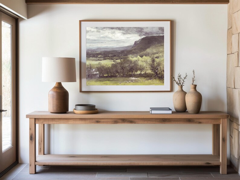 A serene living room setting with a wooden table holding a lamp, ceramic vases, and books, beneath a framed landscape painting of mountains and fields.