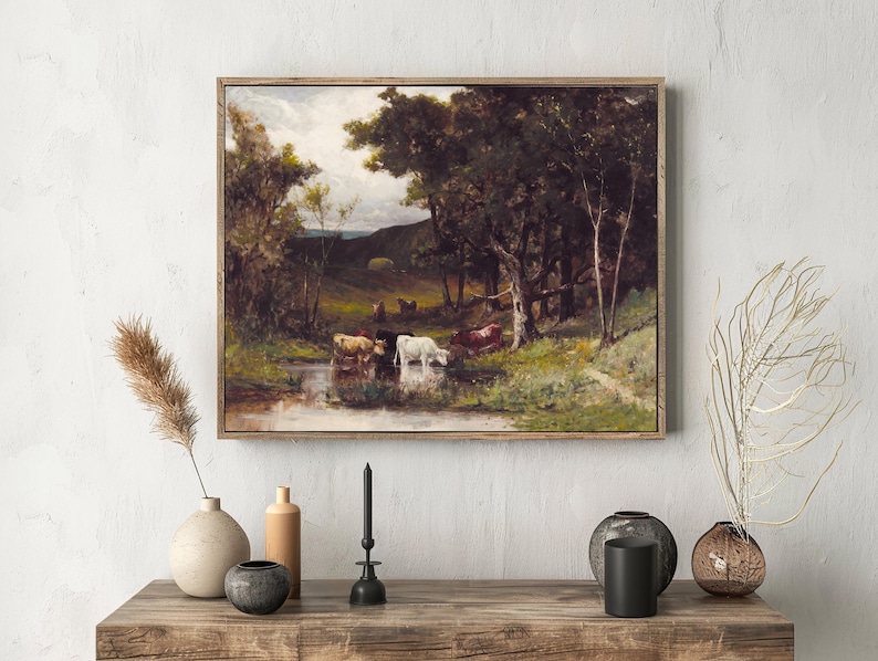 A framed painting of cows by a pond in a lush, forested landscape hangs above a rustic table. Vases with dried plants add a natural touch.