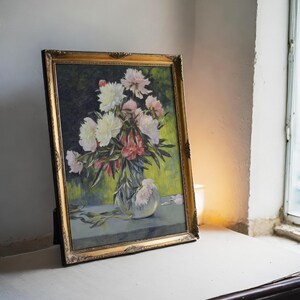 A framed painting of vibrant peonies in a vase, set on a table by a window. Soft light casts a warm glow, highlighting the floral detail and rustic texture.
