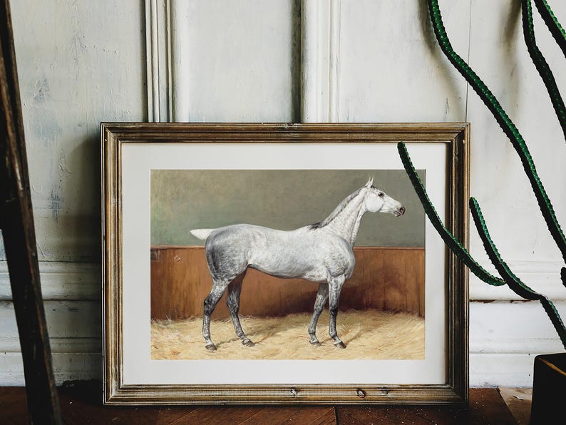 Framed painting of a white horse standing on hay against a brown background, placed on the floor near a cactus. The mood is calm and rustic.