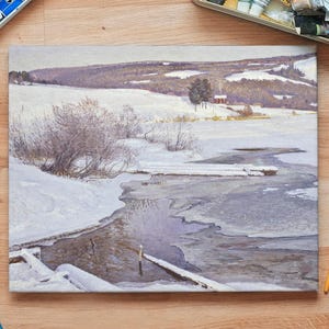 A serene winter landscape painting on a table with art supplies. Snow-covered hills, bare trees, and a frozen river create a peaceful, contemplative tone.