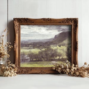 Framed landscape painting with mountains and trees, set against a light wooden wall. Dried flowers in a jar and a bundle add rustic charm.