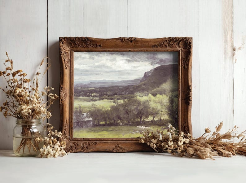 Framed landscape painting with mountains and trees, set against a light wooden wall. Dried flowers in a jar and a bundle add rustic charm.