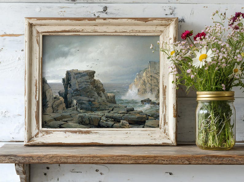 Vintage-style framed painting of a rocky coastline on a wooden shelf, next to a mason jar filled with colorful wildflowers, creating a calm, rustic scene.