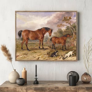 A serene painting of two horses in a rural landscape, with a cloudy sky. Two small dogs play in the grass. The art is framed on a textured white wall, with decorative vases and branches on a wooden shelf below.