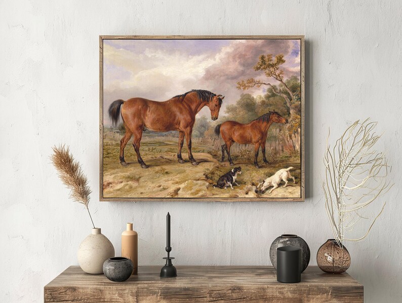 A serene painting of two horses in a rural landscape, with a cloudy sky. Two small dogs play in the grass. The art is framed on a textured white wall, with decorative vases and branches on a wooden shelf below.
