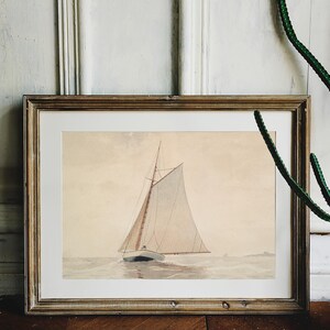 A framed watercolor painting of a sailboat with white sails on a calm body of water. The painting is in a light wood frame.