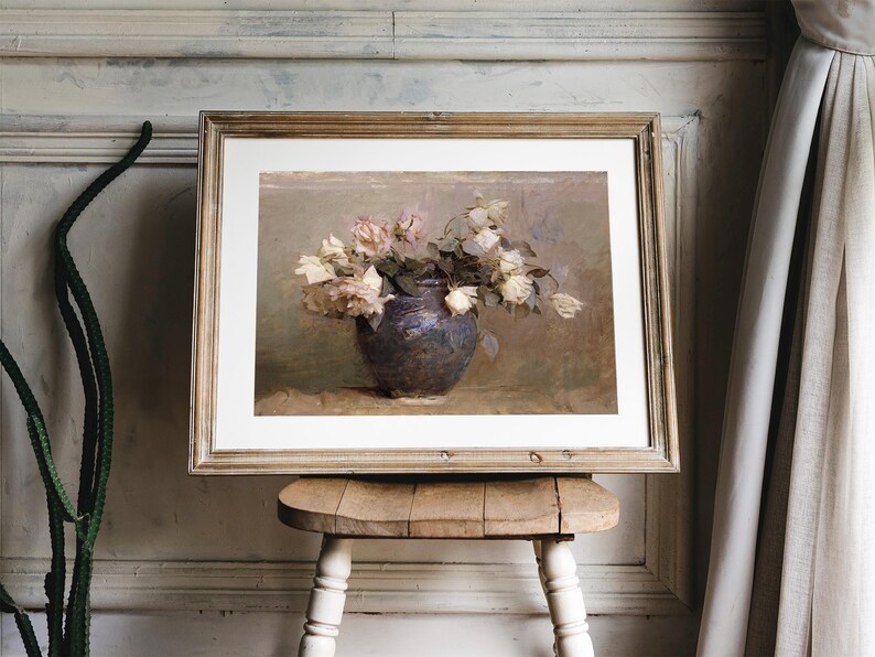 A framed painting of a bouquet of white roses in a dark blue vase. The roses are arranged in a loose, natural style, and the vase is sitting on a table with a light brown tablecloth.