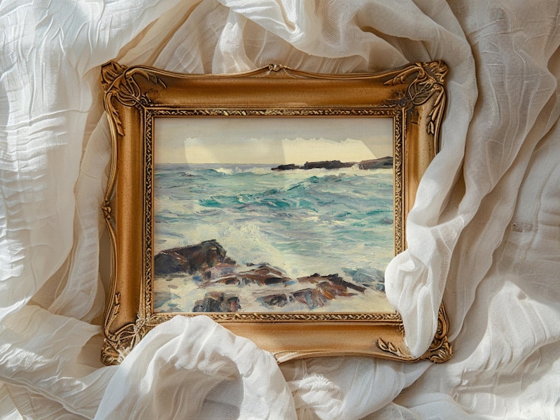 A painting of a turbulent sea with waves crashing against rocks, set in a detailed, ornate gold frame. The frame rests on crumpled, soft white fabric.