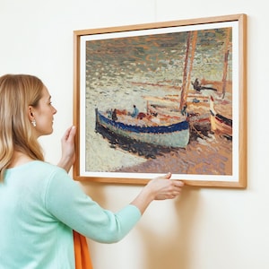 A woman with blonde hair in a light turquoise sweater hangs an impressionist painting of boats on a textured sea. The scene is calm and serene.