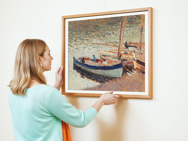 A woman with blonde hair in a light turquoise sweater hangs an impressionist painting of boats on a textured sea. The scene is calm and serene.