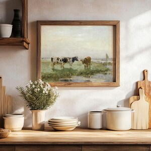 Framed painting of cows in a field above a kitchen counter with stacked dishes, white canisters, a vase with flowers, and wooden cutting boards.
