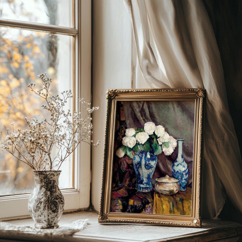 Framed painting of white roses in a blue vase sits on a windowsill beside a vintage vase with dried flowers. Soft light and curtains create a serene mood.