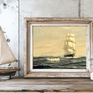 A nautical-themed still life features a model sailboat with beige sails on a wooden surface, a framed painting of a sailing ship on choppy seas in the background, and a glass jar filled with white sand beside the frame. The background consists of distressed, weathered wooden panels in shades of white and gray, enhancing the rustic and vintage ambiance.