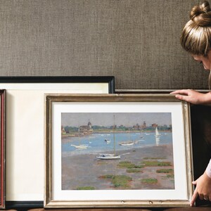 A framed painting of a seascape with sailboats and a small town in the distance. The painting is in a light brown frame with a white mat.