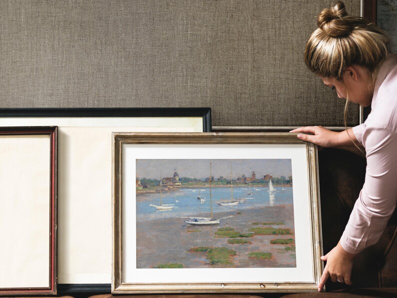 A framed painting of a seascape with sailboats and a small town in the distance. The painting is in a light brown frame with a white mat.