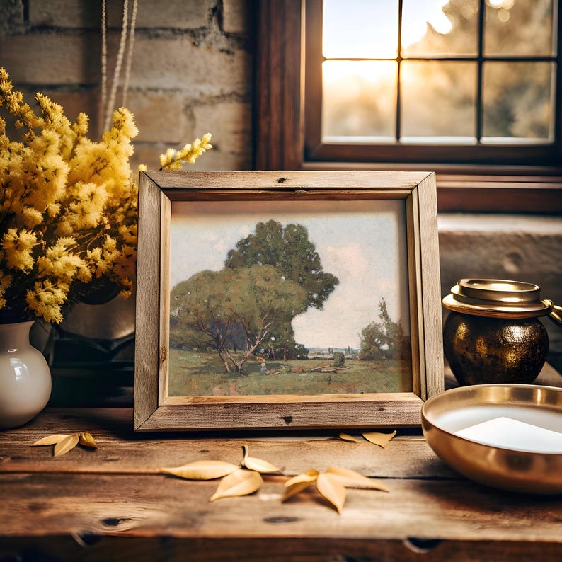 A cozy interior scene featuring a framed landscape painting of a large, leafy tree and open field, placed on a rustic wooden table. To the left, there's a white vase filled with yellow flowers, and to the right, a dark, textured jar with a lid and a shallow gold bowl, all illuminated by warm, natural sunlight coming through a window with wooden framing. There are also some yellow leaves scattered on the table, enhancing the warm, autumnal atmosphere.