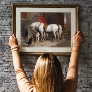 A framed painting of a white horse and two dogs in a stable setting. The horse is wearing a saddle and bridle. The painting is in a wooden frame with a light brown finish.
