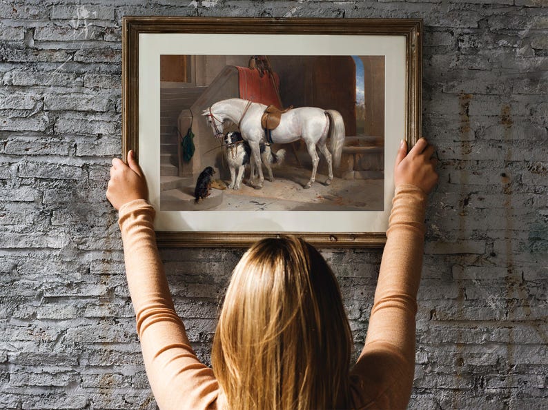 A framed painting of a white horse and two dogs in a stable setting. The horse is wearing a saddle and bridle. The painting is in a wooden frame with a light brown finish.