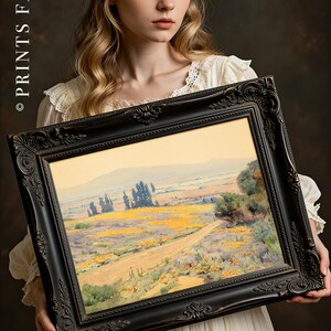 Young woman in a white dress holds an ornate framed painting of a scenic landscape with vibrant yellow fields and distant hills, conveying tranquility.