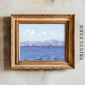 A framed seascape painting depicts a sailboat on a blue ocean with distant hills under a cloudy sky. The frame is ornate and gold, hung on a textured wall.