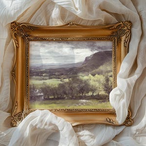 A framed landscape painting rests on crumpled white fabric. The painting showcases a serene countryside with rolling hills, lush greenery, and a cloudy sky.
