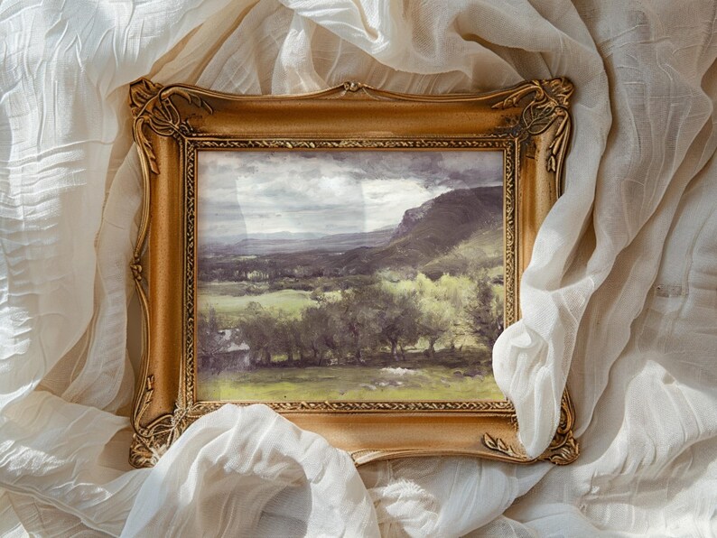 A framed landscape painting rests on crumpled white fabric. The painting showcases a serene countryside with rolling hills, lush greenery, and a cloudy sky.