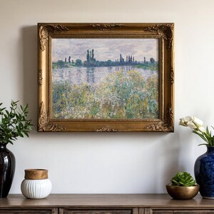 A framed landscape painting with a serene riverside scene hangs on a light wall. Below, a wooden table holds a black vase with greenery, a white jar, a blue vase with white tulips, and a small succulent, creating a calm and elegant atmosphere.