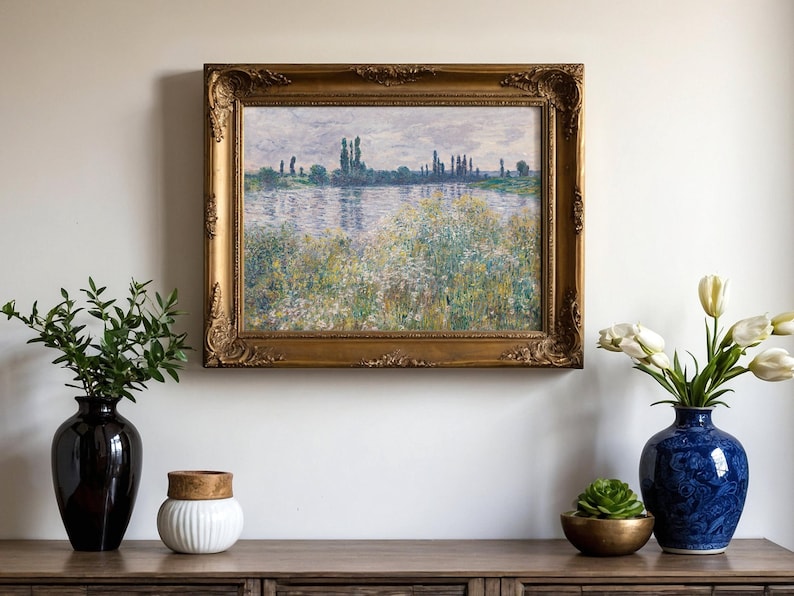 A framed landscape painting with a serene riverside scene hangs on a light wall. Below, a wooden table holds a black vase with greenery, a white jar, a blue vase with white tulips, and a small succulent, creating a calm and elegant atmosphere.