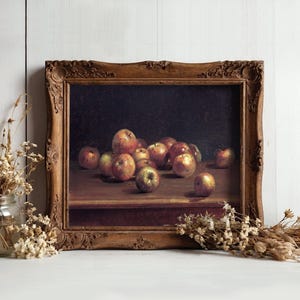 A framed still-life painting of apples on a table, set against a white wooden wall. Dried flowers in vases flank the artwork, adding rustic charm.