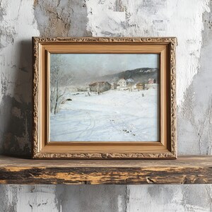 A framed painting of a snowy village scene rests on a rustic wooden shelf against a textured gray wall, evoking a serene and nostalgic winter mood.