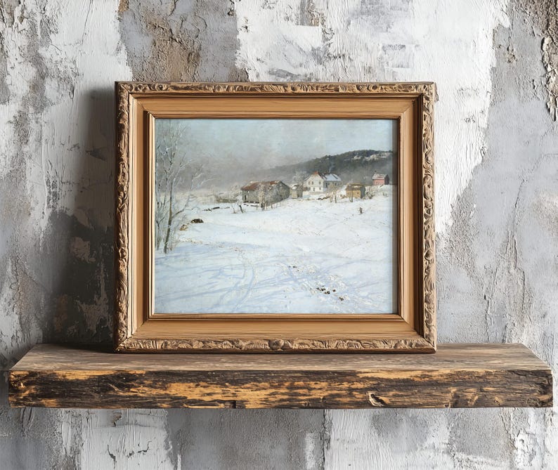 A framed painting of a snowy village scene rests on a rustic wooden shelf against a textured gray wall, evoking a serene and nostalgic winter mood.