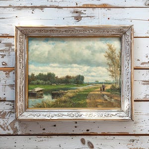 A framed landscape painting on a rustic wooden wall. The scene depicts a serene countryside path near a river under a cloudy sky, conveying tranquility.