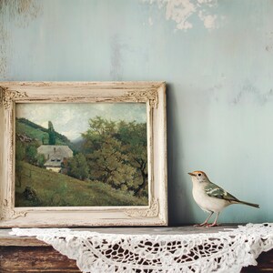A small bird perches on a wooden shelf next to a rustic framed landscape painting. The setting feels vintage and serene with a lace cloth draped over the shelf.