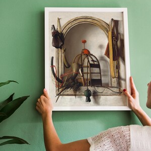 A woman hangs a framed still-life painting on a green wall. The artwork features a birdcage, objects, and an archway, exuding a vintage, serene vibe.