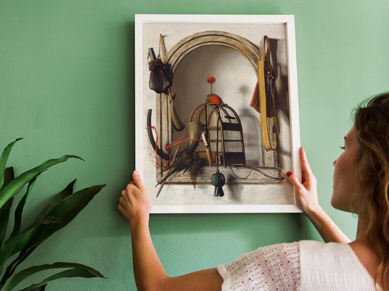 A woman hangs a framed still-life painting on a green wall. The artwork features a birdcage, objects, and an archway, exuding a vintage, serene vibe.
